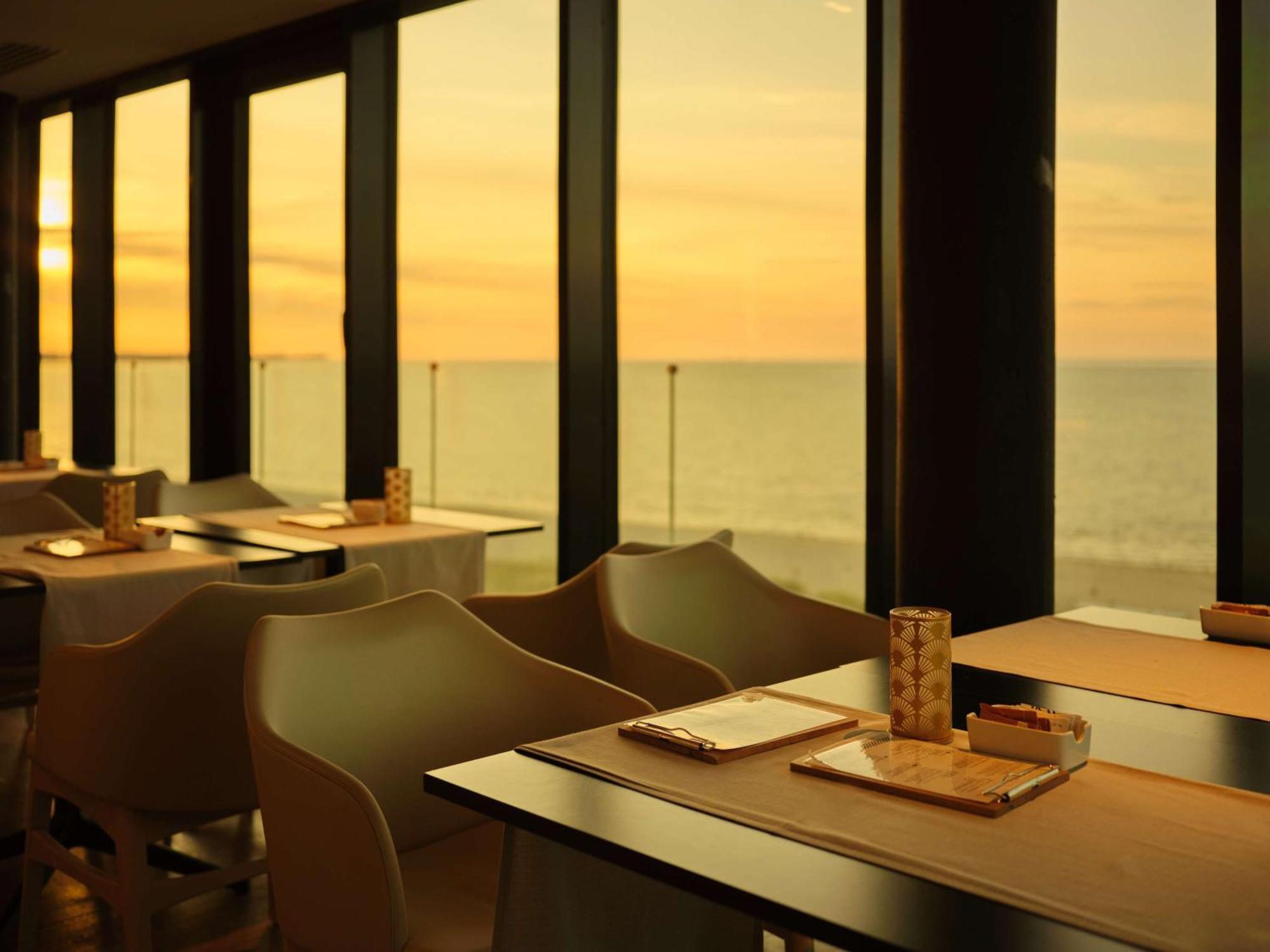 Radisson Blu Resort ชวีโนวิชแจ ภายนอก รูปภาพ The photo shows a restaurant interior with a beautiful view of the ocean during sunset. The large windows allow natural light to flood the space, highlighting the warm colors of the sunset in the sky. There are several tables, each set with placemats
