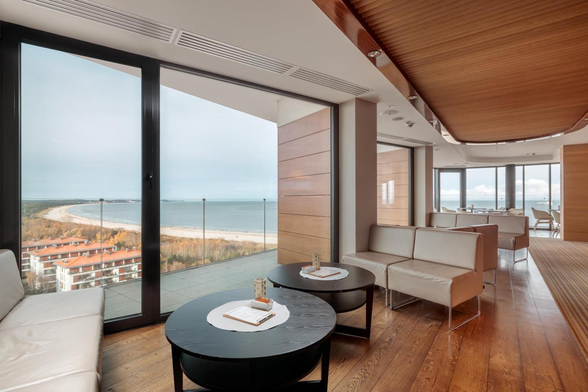 Radisson Blu Resort ชวีโนวิชแจ ภายนอก รูปภาพ The photo shows a modern, elegantly designed interior space featuring large windows that offer a panoramic view of the ocean and coastline. The flooring is wooden, providing a warm atmosphere. There are two round black tables in the foreground with m