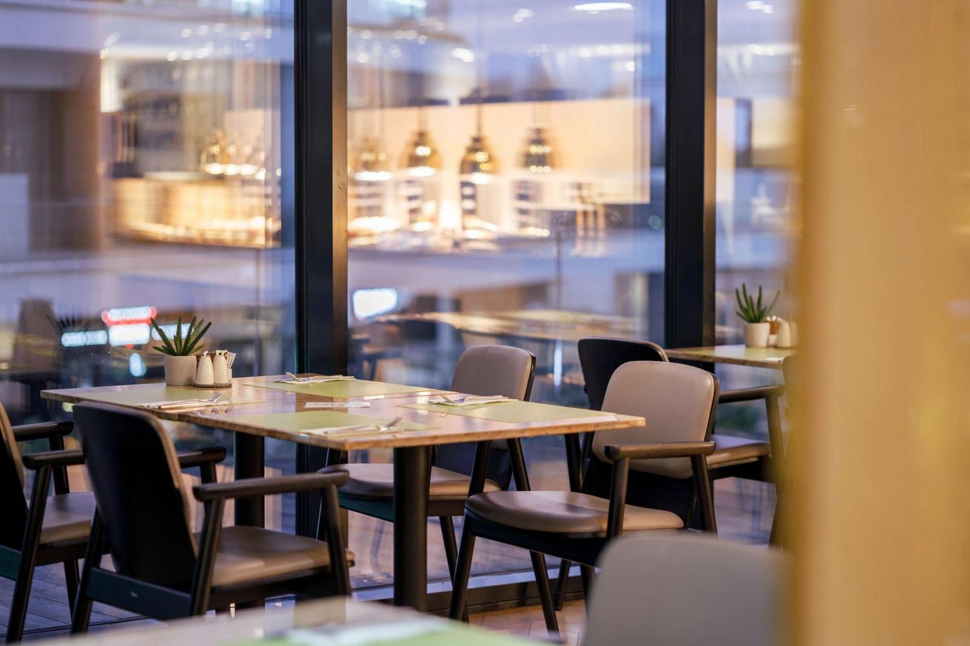 Radisson Blu Resort ชวีโนวิชแจ ภายนอก รูปภาพ The photo shows a modern restaurant interior featuring several tables and chairs arranged under large windows. The tables are set with green placemats and cutlery. There are small potted plants on the tables, adding a touch of decor. In the backgroun