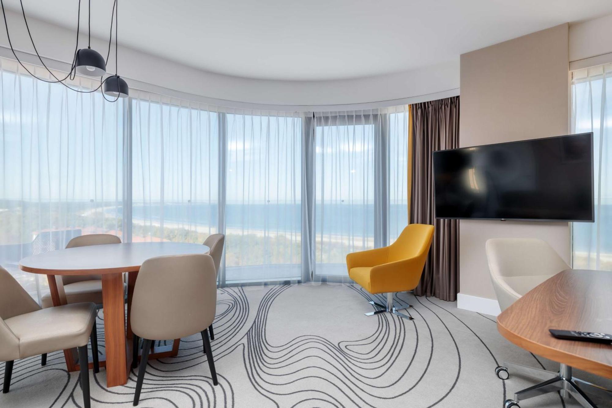 Radisson Blu Resort ชวีโนวิชแจ ภายนอก รูปภาพ The photo shows a modern, stylish interior of a room with large windows that provide a panoramic view. The space features a round dining table surrounded by chairs, including a distinct yellow armchair. There is a flat-screen television mounted on th