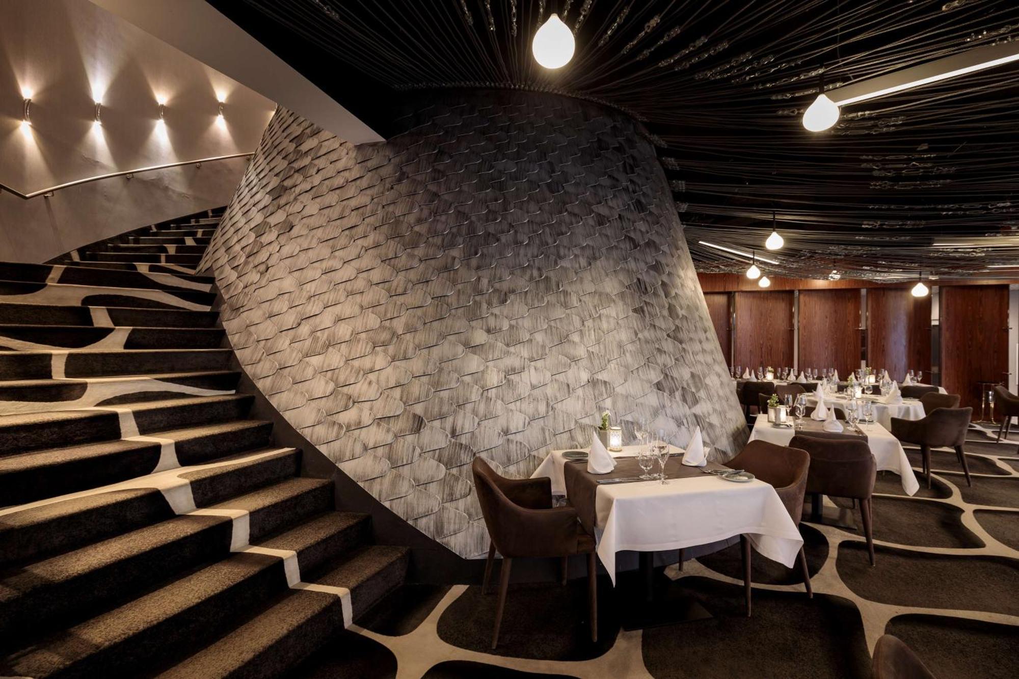 Radisson Blu Resort ชวีโนวิชแจ ภายนอก รูปภาพ The photo shows the interior of a stylish restaurant. In the foreground, there are elegantly set tables with white tablecloths and dark chairs. A unique, curving stone wall adds a distinct architectural feature, and a grand staircase with a textured 