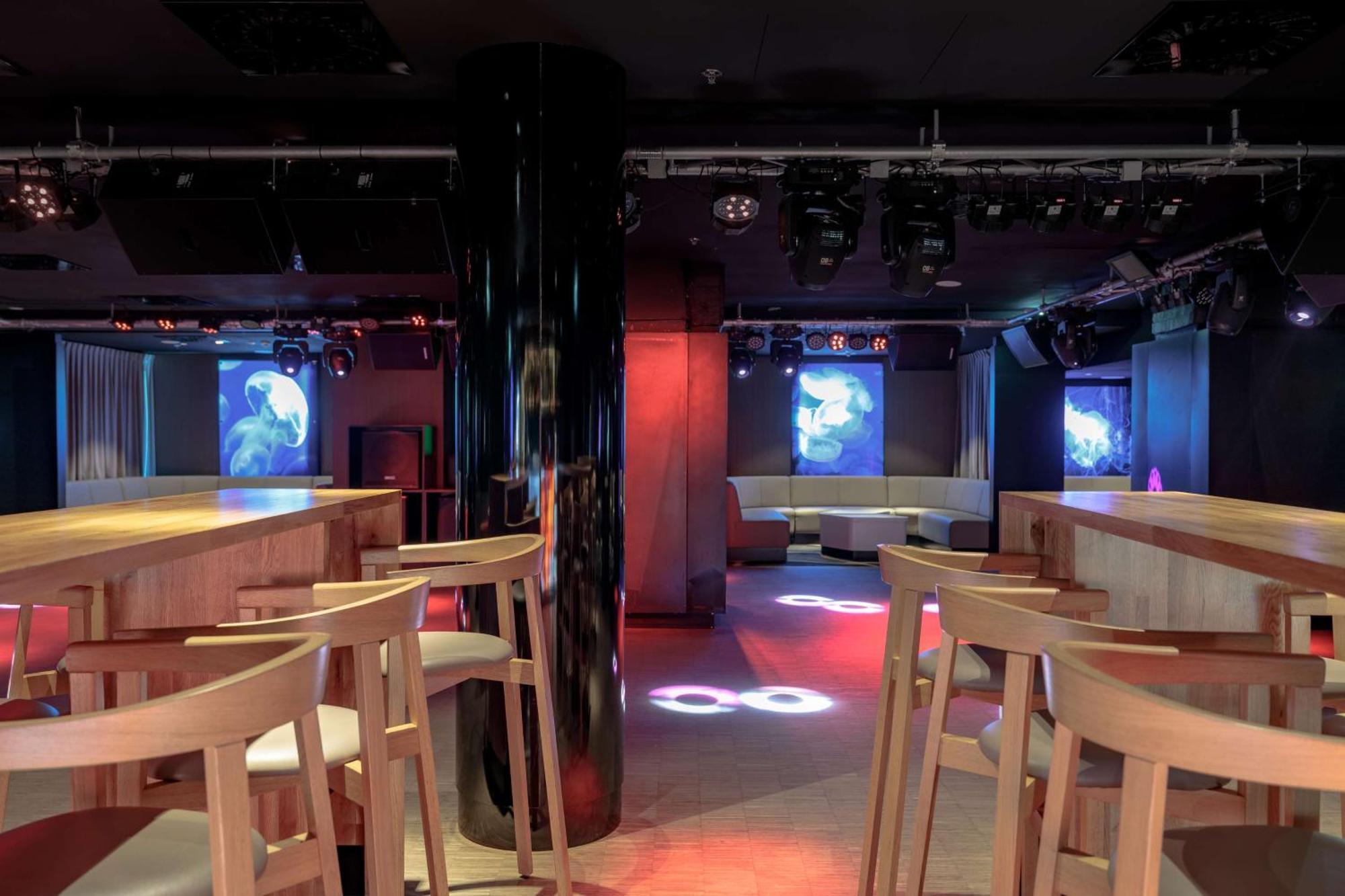 Radisson Blu Resort ชวีโนวิชแจ ภายนอก รูปภาพ The photo shows an interior of a modern venue, likely a bar or club. The space features a central black column and light-colored wooden bar stools arranged around a bar area. On the walls, there are large screens displaying vibrant visuals, possibly 
