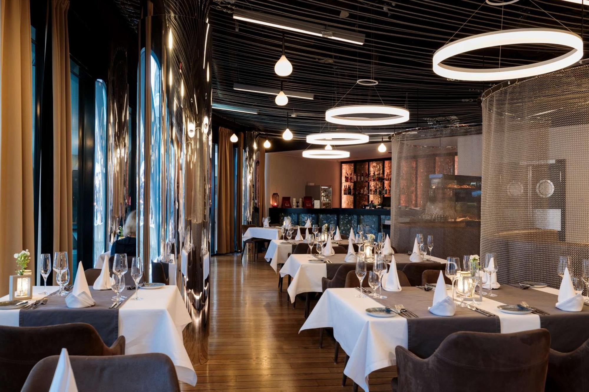 Radisson Blu Resort ชวีโนวิชแจ ภายนอก รูปภาพ The photo shows the interior of a modern restaurant. It features elegantly set tables with white tablecloths and neatly arranged glassware. The design includes a sleek, contemporary aesthetic with dark walls and interesting lighting, including circul