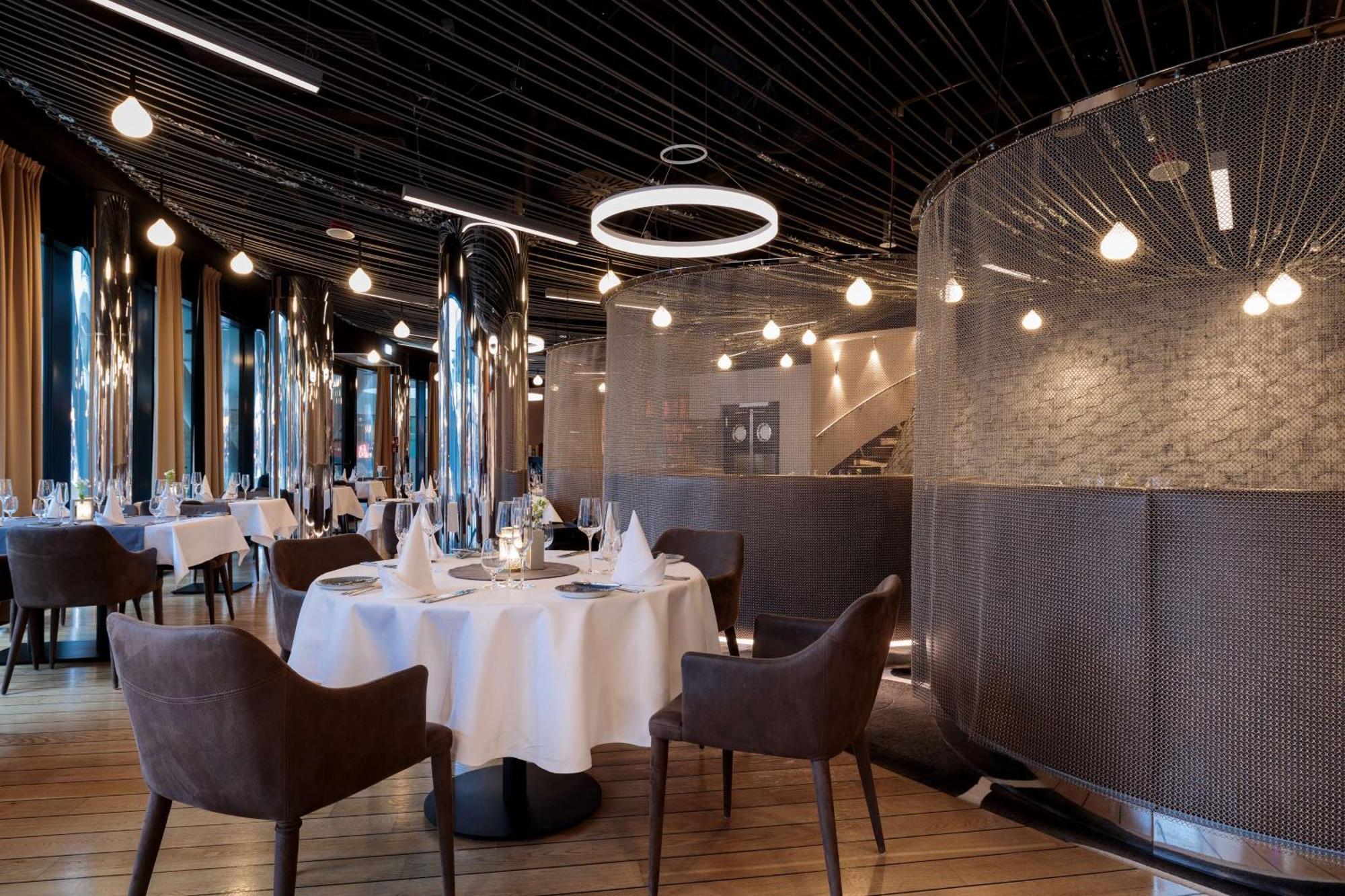 Radisson Blu Resort ชวีโนวิชแจ ภายนอก รูปภาพ The image shows a modern restaurant interior that features a stylish design. There are several round tables with white tablecloths. The seating consists of brown, upholstered chairs. The space is illuminated by various hanging lights, including a dis